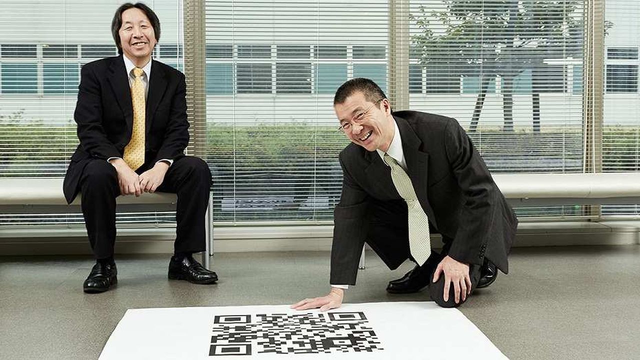 Invention du QR code - Deux ingénieurs japonais de Denso, dont Masahiro Hara assis, présentent fièrement leur invention, le QR code, dans une photo emblématique capturant l'innovation technologique.