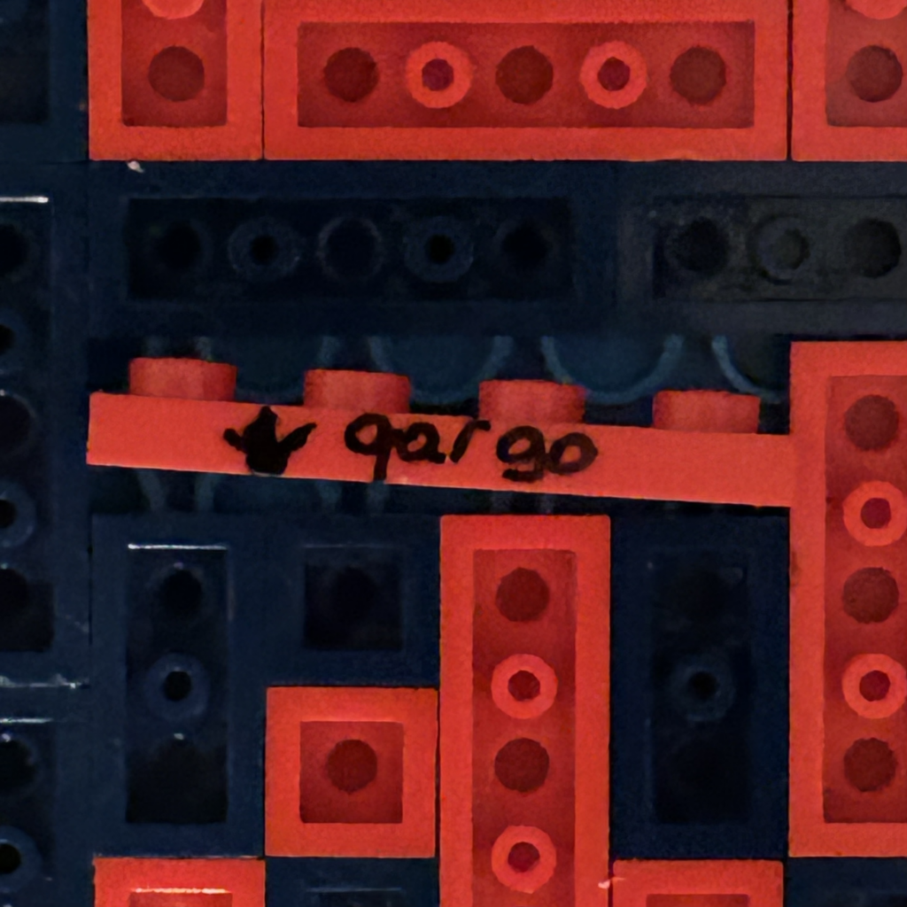 Modèle LEGO plat rouge utilisé par l'artiste qargo comme Easter egg, avec sa signature et un smiley, caché dans chaque œuvre d'art de la collection.